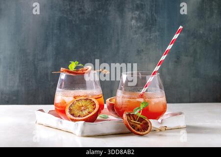 Blood orange iced cocktails in glasses, decorated by slice of oranges and fresh mint, served with retro straw in ceramic tray on white marble table with blue wall at background. Stock Photo