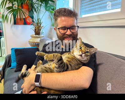 Happy man holding his cat on a couch at home. Companionship, relaxation, and emotional comfort through the bond between human and pet || Model released Stock Photo