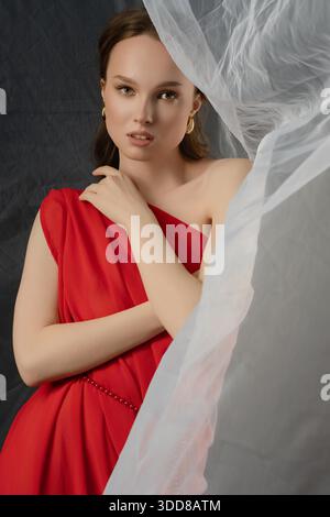 A woman stands elegantly in a studio, donning a flowing red gown. She poses thoughtfully, her expression hinting at various emotions. The backdrop is softly lit, enhancing the mood. || Model released Stock Photo