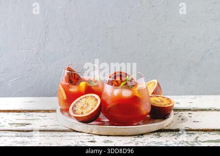 Blood orange iced cocktails in glasses, decorated by slice of oranges and fresh mint, served in ceramic tray on white wooden table with grey wall at background. Stock Photo