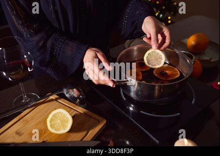 A warm, festive moment of a woman in a dark, elegant dress preparing mulled wine with citrus slices on the stove, candlelight, and a glass of wine nea Stock Photo
