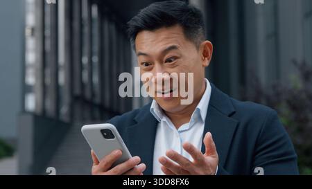 Happy shocked Asian business man looking at phone amazed Korean businessman in city 40s male Chinese Japanese executive feeling surprised wonder Stock Photo