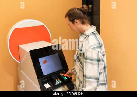 Young man with hair bun inserting credit card into ATM machine to withdraw cash at bank. High quality photo Stock Photo