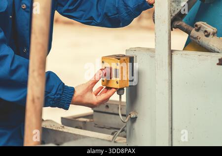 Close-up of worker s hand in blue coveralls pressing control button on industrial machine at manufacturing facility. Industrial safety Stock Photo