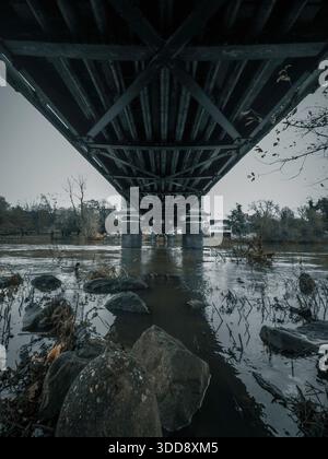 A symmetrical view beneath a steel bridge with concrete pillars standing in a calm river. Industrial architecture, water reflections, and an urban env Stock Photo