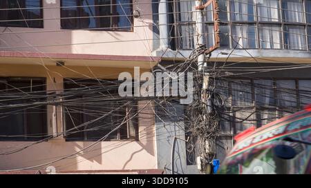 Tangled electrical cables clustered around a power pole in front of an unsanitary, weathered building, illustrating urban infrastructure challenges in Stock Photo