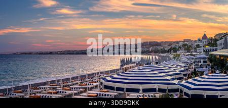 Blue and white striped umbrellas on a pebbled beach, panorama of a golden sunset over Mediterranean Sea along the Promenade des Anglais, Nice, France Stock Photo