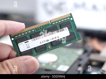 Close-up of fingers holding a green PCB RAM stick against a blurred workshop background. Visual for computer technicians, hardware inspection, and par Stock Photo