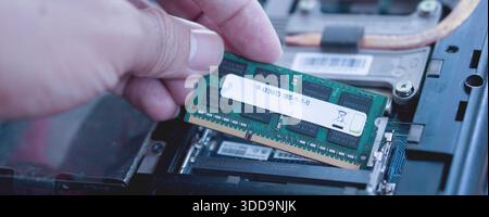 A technician's hand carefully inserting a green RAM stick into the laptop's memory slot. Visualizes hardware maintenance and performance tuning. Stock Photo