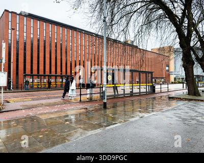 Manchester, UK - December 06, 2025: Pedestrians in coats walk by a red brick Kilburn Building on a wet urban street in Manchester. Stock Photo