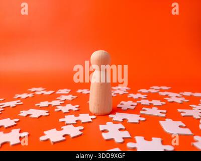 Wooden peg doll surrounded by white jigsaw puzzle pieces on an orange background. Stock Photo