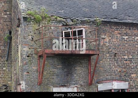 derelict window located in an old disused building. bricks and roof are falling and the metal balcony is rusty. Vegetation is growing out of brickwork Stock Photo