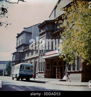 Slaviansk, Donetsk region, Ukrainian SSR, Soviet Union, 1980s. An atmospheric archival color photograph of the Palace of Culture of the Ceramic Combine located on Lenin Street (now Universitetskaya). The historic grey building features arched windows, a balcony, and a red banner above the wooden entrance doors. A blue and white Soviet service bus (KAvZ model) with Donetsk license plates is parked on the street. Yellow autumn leaves frame the right side of the image. A scene of urban architecture and public transport in the peaceful Donbas. Stock Photo