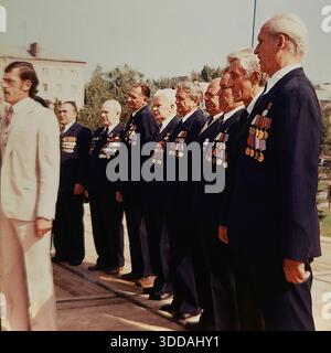 Slavyansk, Ukraine, USSR, 1980s. Performance of the War Veterans Choir of the SlavTyazhMash heavy engineering plant on October Revolution Square (now Soborna Square). A soloist in a light suit sings in front of a microphone, while a row of elderly men in dark suits adorned with numerous WWII military medals and orders stands behind him. This archival image captures the patriotic culture and honoring of war heroes in the industrial Donbas region. Stock Photo
