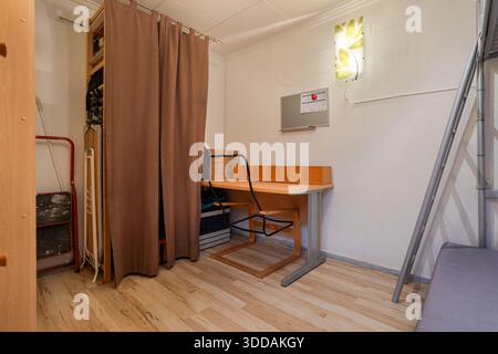 Small study nook in bedroom featuring desk, chair, shelving behind curtain, and soft wall lamp Stock Photo