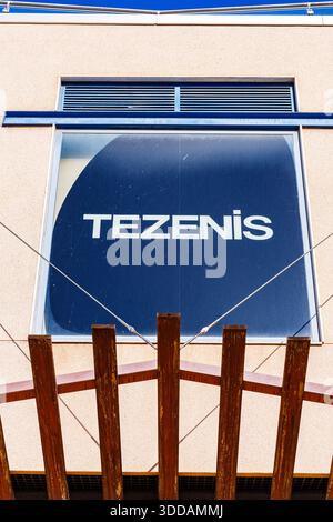 The Tezenis brand logo displayed on a dark blue window panel of a shopping center building, framed by wooden beams and industrial cables. Stock Photo
