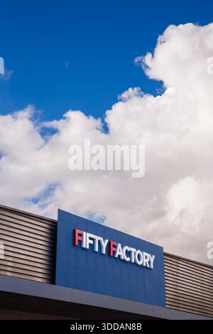 The clean, modern signage of the Fifty Factory outlet store set against a bright blue sky and large white cumulus clouds. Stock Photo