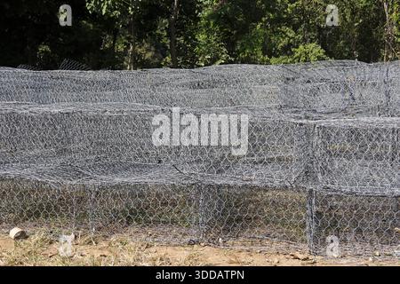 Solid protective gabion wall made of galvanized wire mesh cage structure filled with rock stone standing strong near green forest in peaceful outdoor Stock Photo