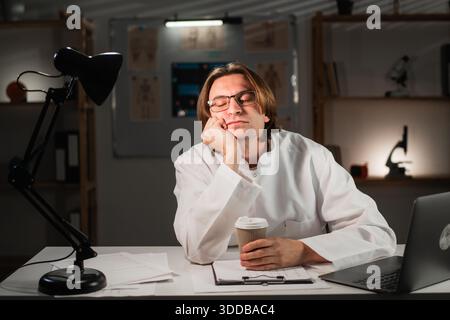 doctor in uniform sleeping tired posing with hands sitting at table during night shift in clinic. Stock Photo