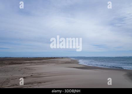 Wide stretch of undisturbed sand meets gentle waves under a soft, cloud-filled sky at Plage Napoleon, Port Saint Louis du Rhone. Subtle patterns in the sand and muted daylight create a serene coastal scene. Stock Photo