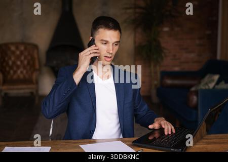 Handsome caucasian manager sitting at office desk in front laptop hold mobile phone, make pleasant business or informal call. Successful businessman looking at financial statistics shown on documents || Model released Stock Photo