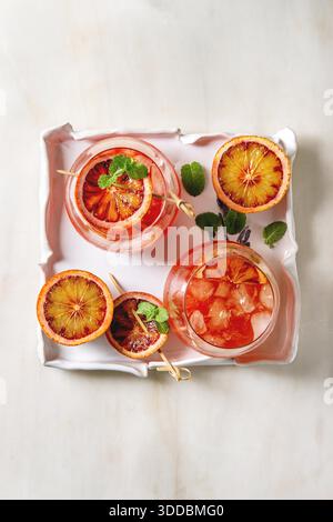Blood orange iced cocktails in glasses, decorated by slice of oranges and fresh mint on skewers, served in ceramic tray over white marble background. Flat lay, space. Stock Photo