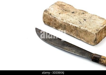 An old, rusty machete lays on a white surface next to a big thick whetstone, porous texture. Both objects show signs of weathering and wear, suggestin Stock Photo