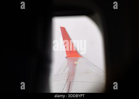 View of an aircraft wing with a prominent red winglet seen through an airplane window frame. The wing's metallic surface is visible against a bright, Stock Photo