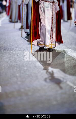 A close-up focus on the lower half of a penitent’s white silk robe and red velvet cape, highlighting the golden trim and the shadow cast on the street Stock Photo