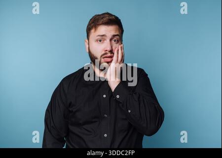 A man in a black shirt looks surprised, holding his cheek. Stock Photo