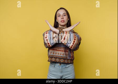 Woman in patterned sweater making an X with arms on yellow background. Stock Photo