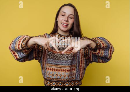 Smiling woman in a patterned sweater making a heart shape with her hands against a yellow background. Stock Photo