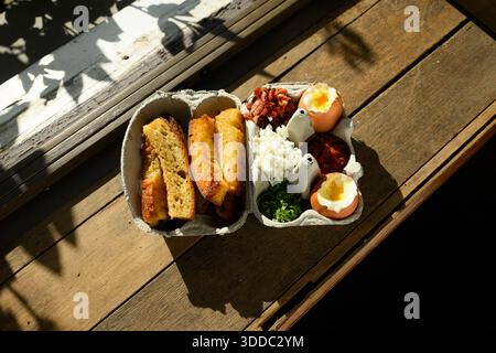 A tray of food is on a wooden table. The tray contains a sandwich, an egg, and some vegetables. The sandwich is cut in half and placed on the table. T Stock Photo