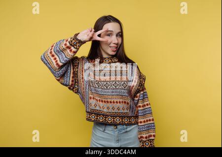 Woman in patterned sweater making a peace sign on yellow background. Stock Photo