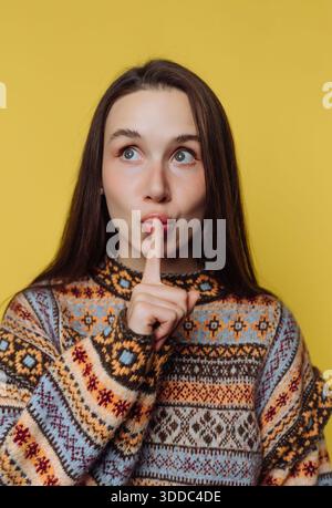 Woman in patterned sweater making a shushing gesture against a yellow background. Stock Photo