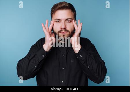 Man in black shirt touching face with both hands against blue background. Stock Photo