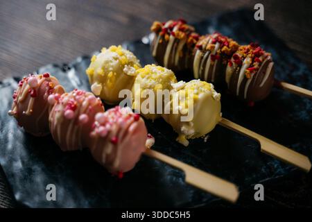 Three different colored desserts with white frosting and sprinkles on top. The desserts are arranged on a black plate with wooden sticks. The desserts Stock Photo