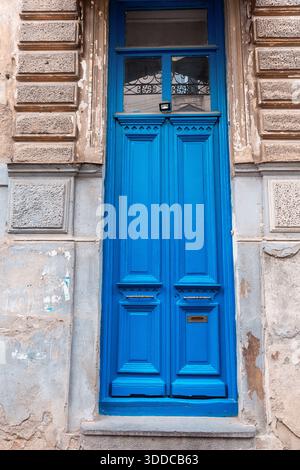 Bright blue ornate wooden entrance door with a glass transom window and mail slot on an old, weathered building wall Stock Photo