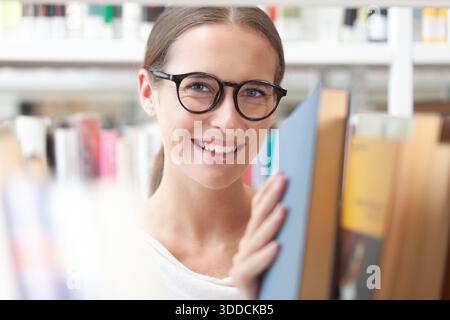 Woman wearing glasses choosing a book in a library or book shop, seen through shelves with books in the foreground. Concept of education, learning, re Stock Photo