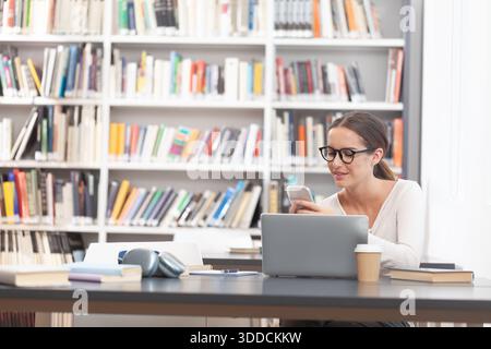 Young woman using smartphone sitting at desk with laptop, female student studying in a college library, Concept of education, studying, research, know Stock Photo