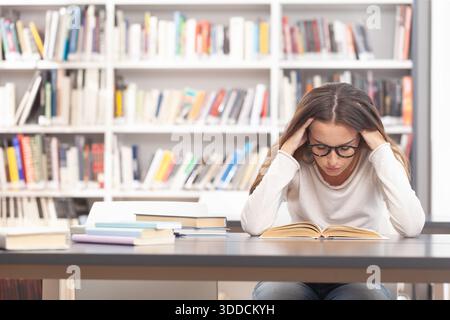 Young female university student sitting at a desk in a library, holding her head while reading a textbook. Academic stress, exam preparation, and ment Stock Photo