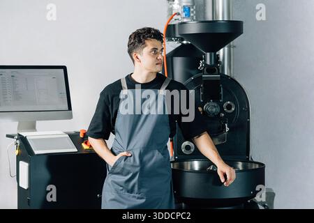 Confident coffee shop worker in an apron leaning roasting machine. Young coffee roasting machine operator looking away. Stock Photo