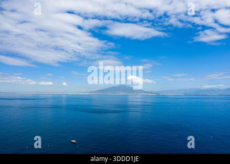 Clear blue waters of the Bay of Naples stretch toward the distant silhouette of Mount Vesuvius beneath a sky filled with scattered white clouds. A small boat floats in the foreground, adding scale to this iconic Italian coastal scene. Stock Photo
