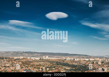 Lenticular cloud forming in the vivid blue sky above a sprawling autumn cityscape, with distant mountains along the horizon Stock Photo