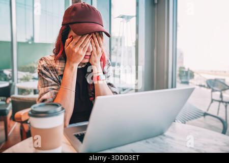 Young woman wearing a cap and plaid shirt, covering her face with hands, feeling overworked and stressed while using a laptop Stock Photo