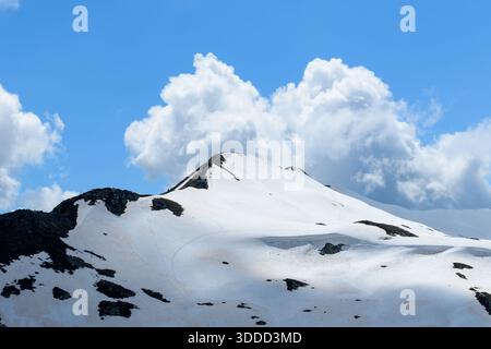 Aiguillette des Houches displays a smooth snow-covered slope with scattered dark rocks, set against a backdrop of vibrant blue sky and large, billowing cumulus clouds. Stock Photo