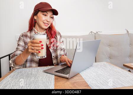 Young woman freelancing in a cafe, sitting at a wooden table with a laptop, typing and drinking a coffee latte Stock Photo
