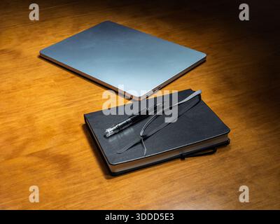 A professional setup featuring a fountain pen resting horizontally on a closed black notebook with a sleek laptop nearby on a wooden surface. Stock Photo