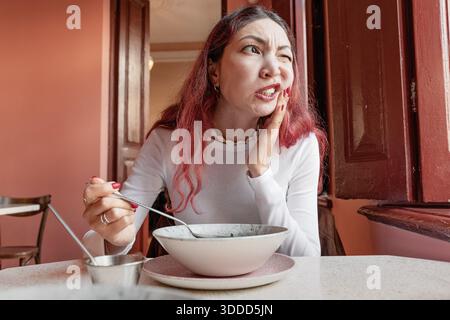 Woman having an intense tooth pain, holding her painful cheek, grimacing, experiencing discomfort from dental problems while eating Stock Photo