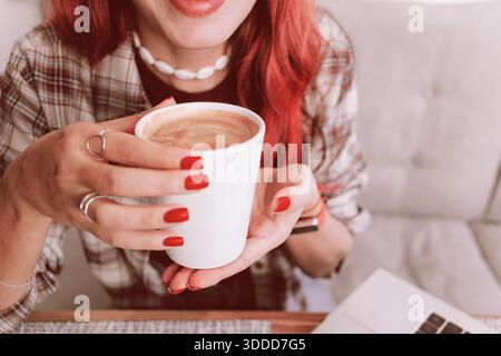 Red haired woman with red manicure holding a warm white coffee mug by her laptop, relaxing in a cozy work from home moment Stock Photo